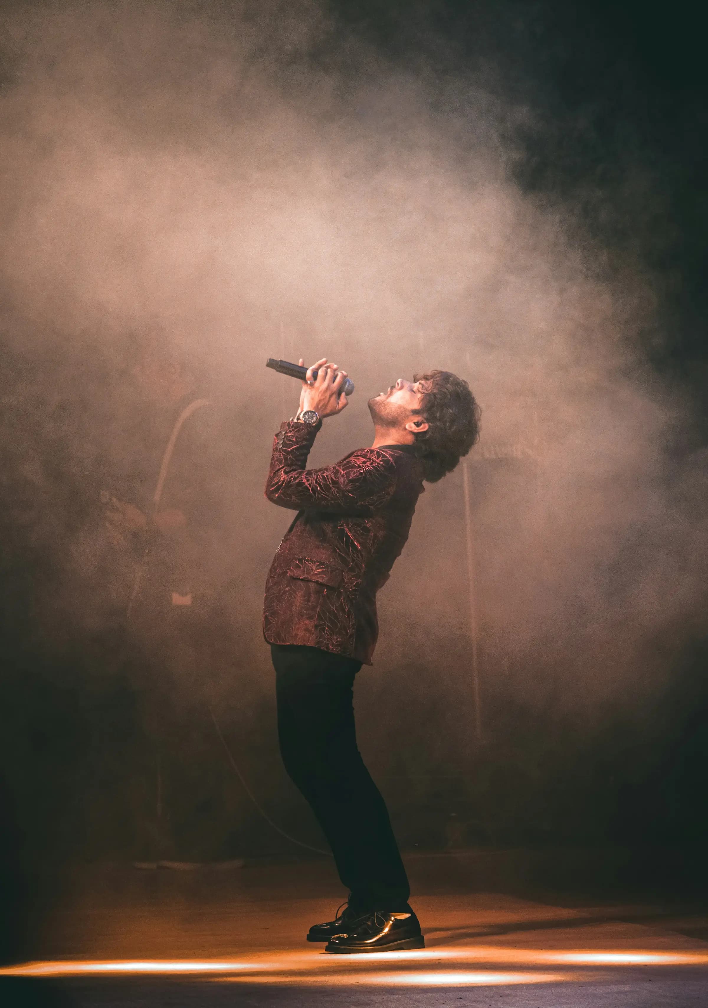 A singer leans back into the haze, microphone raised, under warm stage light